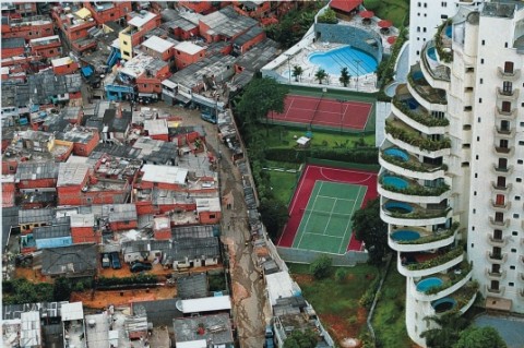 A fence separates a luxury apartment building with green space recreational areas next to a crowd of low income houses