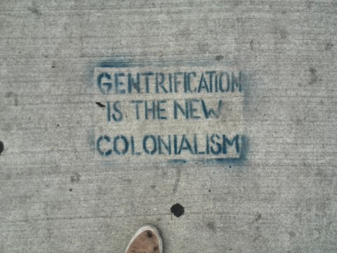 The words "Gentrification is the new colonialism" spray painted onto sidewalk.