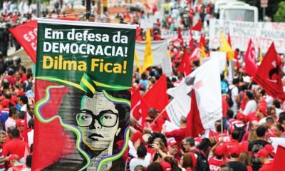 A group of people hold red flags and protest. A sign saying "In defense of DEMOCRACY! Dilma Stays!" in Portuguese